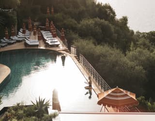 Early light brushes the calm ocean and still waters of the hotel pool, where a lone guest sits at the edge, seen from above.