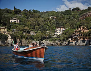 A couple explore Portofino Bay in a wood hulled motor boat. Behind them, Hotel Splendido emerges from the trees