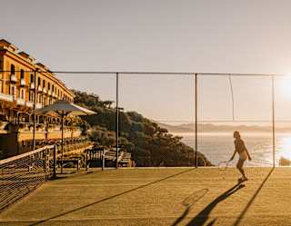 A person plays tennis on an outdoor court at sunset, with the sun low over the sea. Nearby, a yellow building with balconies overlooks the court and water, and trees line the coast.