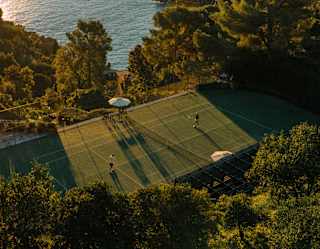 Aerial view of a tennis court surrounded by lush trees and hills, overlooking a calm sea during sunset. Four players are on the court, whilst long shadows stretch across the ground.
