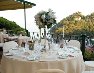 A centrepiece of a flower-filled silver candelabra adorns a white wedding table. Over the balcony, sailboats bob in the harbour