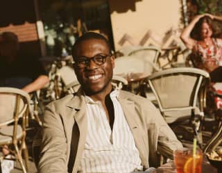 A young man in cream linen blazer smiles at the camera from a bar terrace. A glass of Negroni is on the table in front of him