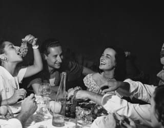 A group of people sit laughing, their table full of drinks and fruit. A young woman dangles a bunch of grapes into her mouth