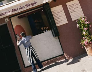 Angled image of a dark-haired waitress standing at the hatch of Gelateria San Giorgio in a striped apron, seen from behind.