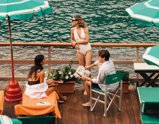 Young people in bathing suits relax on a large balcony with green parasols as the sparkling emerald sea extends far beyond