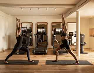 Two people practise yoga on mats in a gym, both in a warrior pose with arms raised. Behind them are three treadmills and large mirrors on the wall. The room has wooden floors and a bright, minimal design.