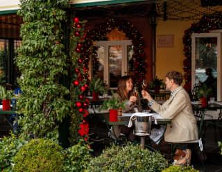 A man and woman raise glasses as they celebrate the season at a table for two on Dav Mare's terrace, seen from outside.