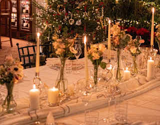 A table, decorated for a feast sparkles with glasses, candles, flowers vases and a twinkling Christmas tree behind.
