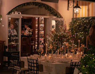 View of a dining room glowing and twinkling with festive decorations, with focus on a table filled with glasses and candles.