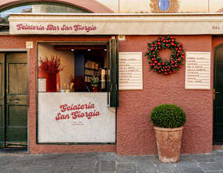 A fir and red-bauble wreath hangs on the terracotta wall between the open hatch and entrance door of Gelateria San Giorgio.