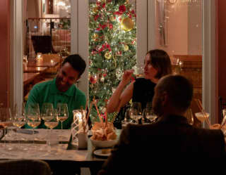 A Christmas tree glows behind glass doors, in front of which a woman sips wine with two male friends at a festive table.