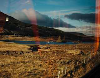 The train passes golden-grassed plains around a small lock, seen through a window that reflects hints of the interior.