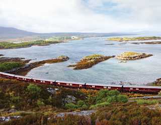 Burgundy train carriages curving alongside a rugged loch dotted with little islands