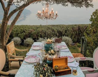 The garden table is set for a celebration beneath a chandelier suspended from an olive tree, with breathtaking coastal views.