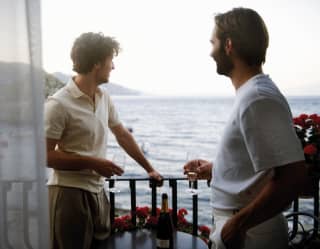 Looking from suite doors on to a balcony where two men gaze out at the Ionian coastline, holding glasses of sparkling wine.