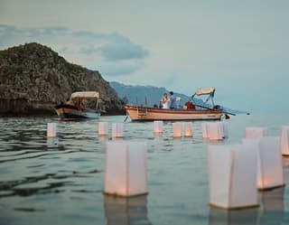 White paper box lanterns float on the calm evening sea as a young couple in a boat clink glasses on their romantic getaway