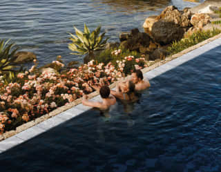 Three swimmers rest at the edge of the pool, gazing out past a small rock garden beach and nearby sea stacks to the ocean.