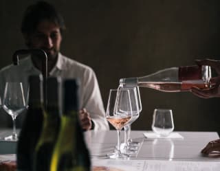 A man sits at a large table with several wine glasses and bottles of wine. A rosé is decanted into three glasses, ready for tasting