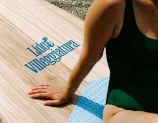 A woman in a dark green swimming costume and sunglasses sits on a wood-effect paddleboard branded with Lido Villeggiatura.