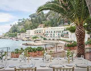 A long table is exquisitely set for lunch on the dining terrace amid palm trees and looking across the Bay at the sweep of hotel