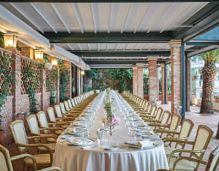 Long banquet table set for a wedding under a lamplit pergola surrounded by vines