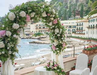 A wedding arch of green foliage with pink and white flowers frames the view of the Bay before a congregation of white chairs