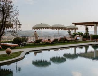 Seen across the still pool waters, guests relax on loungers beneath pretty fringed parasols, absorbing the spectacular views.