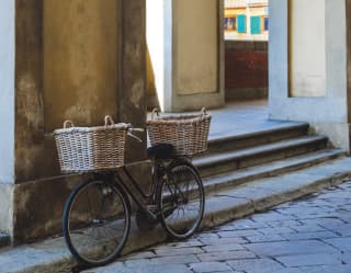 A traditional black bicycle with front and back wicker baskets rests against a wall in the shadows of a quiet Florence lane.