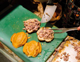 A chef with a large knife and prong builds two crusty paninis with just-roasted pork during a foodie tour, seen from above.