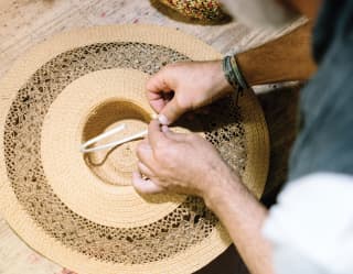 Renowned milliner Antonio Gatto threads a chin strap on a delicate straw hat during a demonstration, seen from above.