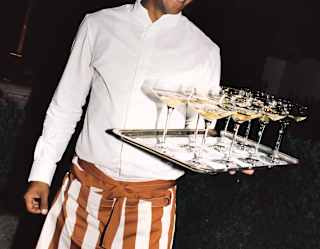A smiling waiter wearing a white shirt and striped apron holds a tray with several glasses of champagne at an evening event outdoors.