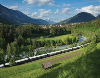 Blue and cream train carriages curving across a sunny Alpine valley