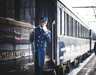 Train guard in a blue uniform blowing a whistle next to train carriages