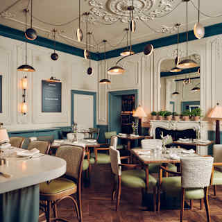 Elegant restaurant interior with ornate white walls, gold accents, and wooden herringbone floors. Round tables are set for dining, with cushioned chairs and soft lighting from modern hanging lamps and table lamps.