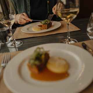 View over a soft-focus plate of food to a guest cutting into meat in a dining table detail with two glasses of white wine.