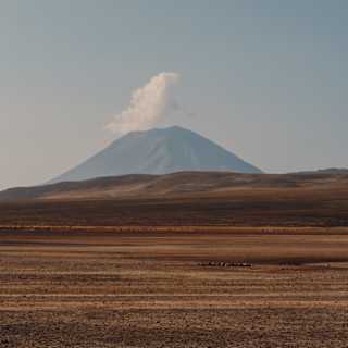 A distant volcano with a plume of white smoke rises above a barren, brown landscape under a clear blue sky.