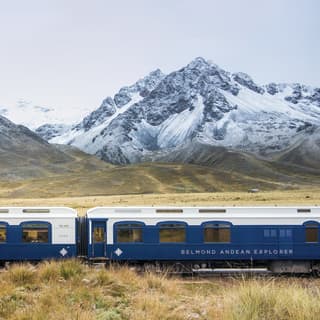 Two white and navy train carriages before snow coated Andean mountains