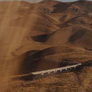 The Andean Explorer train journeys along a snaking track through undulating hills, seen from above through golden sun haze.