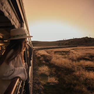A female passenger in a brimmed hat leans on the railings of the observation deck as the sun sets over the golden plains.