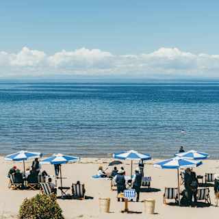 People relax on a sandy beach under blue and white parasols, facing a calm, expansive body of water with distant mountains and clouds on the horizon under a clear blue sky.
