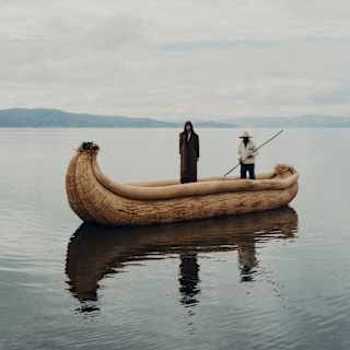 A figure in dark clothes stands in a banana-shaped vessel of bound totora reeds on Lake Titicaca, steered by a local boatman.