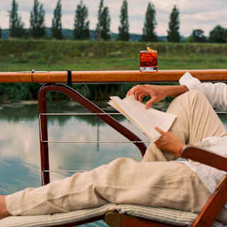 A man in casual clothes reclines in a chair, reading a book with a Negroni-style drink next to him on a deck's wooden rail.