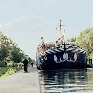 Water-level view of the Amaryllis on a narrow section of the Canal du Centre, about to be moored by a man holding a rope.