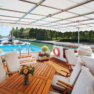 Wooden table surrounded by white fabric chairs on the top deck of a luxury barge