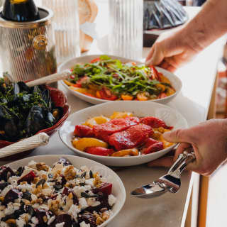 A guest reaches for bowls of roasted capsicums and bright tomato salad at a buffet table with dishes, bread and champagne.