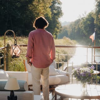 A man in a pink shirt and white trousers with dark hair watches the sun dance on the water from the deck, seen from behind.