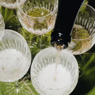 Effervescent champagne is poured evenly into six crystal glasses on the green lounge table, seen close-up from above.