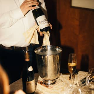 At a table laid for dining, with wineglasses and gold-plated cutlery, a waiter shows guests the label on a bottle of wine.