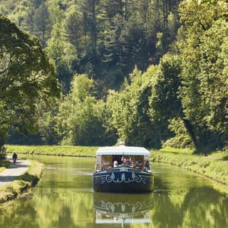 The Fleur de Lys cruises up the serene Ouche river, bordered by big trees and green banks, where a cyclist enjoys the route.