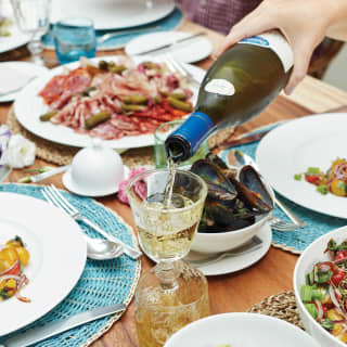 Mão servindo vinho branco em uma taça sobre uma mesa repleta de pratos da culinária francesa